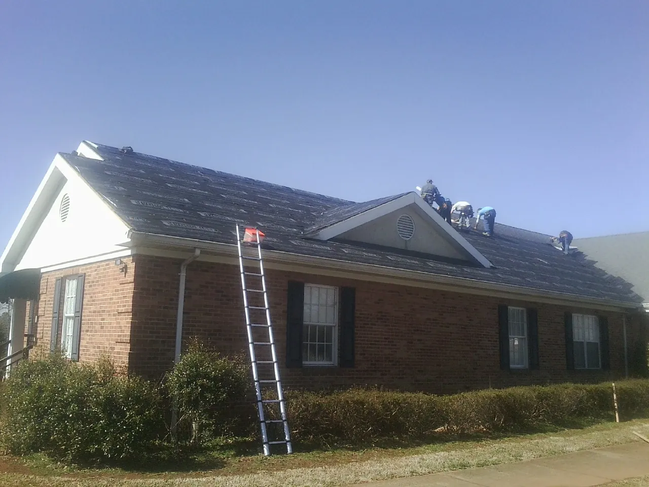 Crew tearing shingles off a brick ranch roof with a ladder set — Metal Roof Repair work by our crew in Royalston