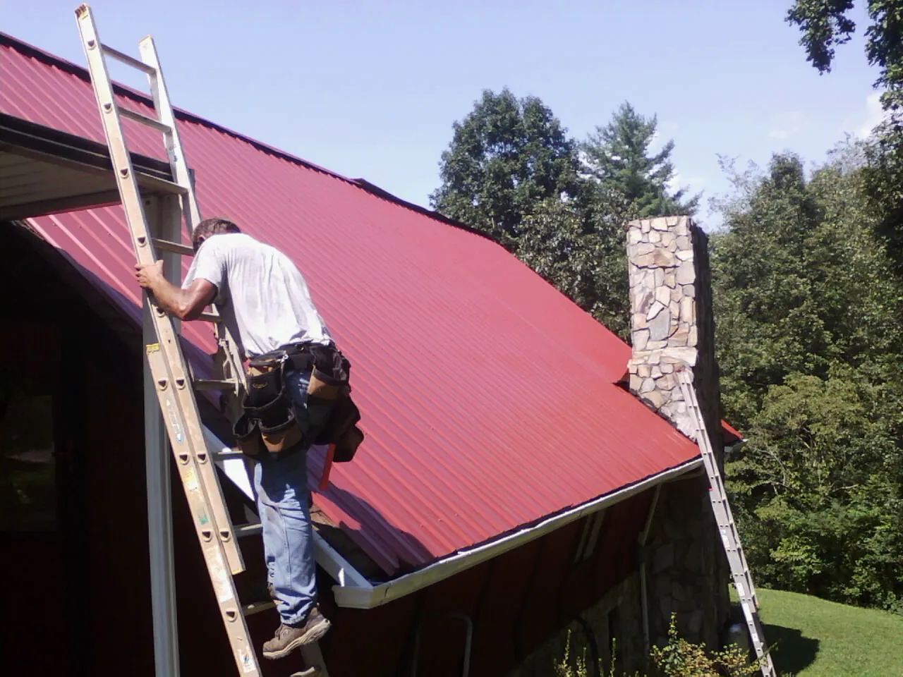 Roofer on a ladder beside a red metal roof and stone chimney — Soffit & Fascia Repair by Roofing Brothers in Royalston